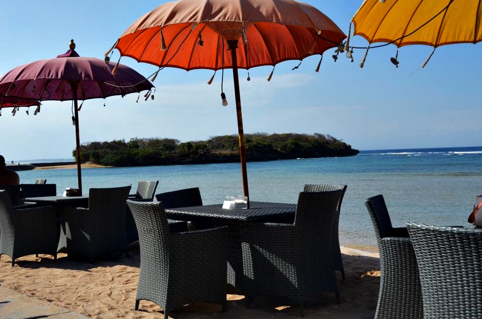 Let's cheer for Bali and the ocean here. Beach bar tables with bright parasols and ocean behind